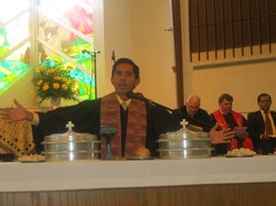 A reverend in vestments before a church, performing communion with his hands held out.